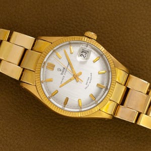 May include: A gold-toned wristwatch with a silver face and a date display at the 3 o'clock position. The watch features gold hands and hour markers, and a gold band. The watch is a Tudor Prince Oysterdate self-winding model.