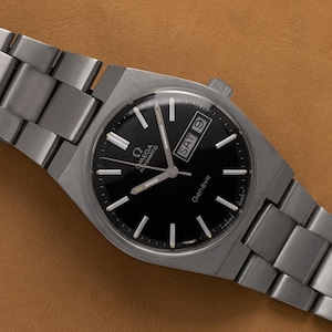 Pode incluir: Um relógio de pulso automático Omega Genève em aço inoxidável com mostrador preto, índices de bastão prateados e janela de dia e data na posição das três horas. O relógio possui uma pulseira de elos de metal escovado e caixa redonda.