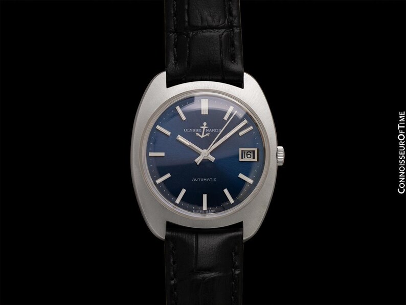 May include: A vintage Ulysse Nardin automatic wristwatch with a dark blue dial and silver hands and markers. The watch has a date window at the 3 o'clock position and is set on a black leather strap. The watch case is silver and has a unique shape.