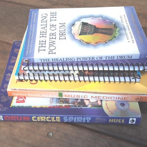 May include: A stack of books on a wooden surface. The titles include "The Healing Power of the Drum," "Music Medicine," and "Drum Circle Spirit." The books are in various colors, including blue, yellow, and orange. A spiral-bound notebook is also visible.