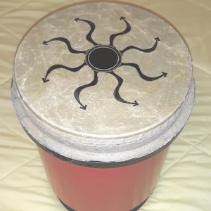 May include: A red drum with a circular top featuring a black sunburst design. The drum's body is red, and the top is a light tan color. The drum is a percussion instrument.
