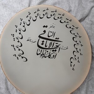 May include: A round, off-white decorative plate with black calligraphy. The text is arranged in a circular pattern around the edge and in the center. The plate has a light brown rim and is set against a gray background.
