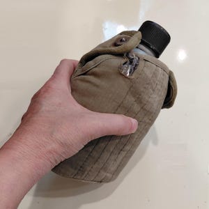 May include: An olive green military-style canteen with a black cap. The canteen is encased in a matching canvas cover with a button closure. The canteen is held in a hand.