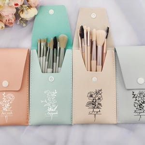 May include: Four personalized makeup brush holders with floral designs and the name "Aeyah" printed on each. The holders are in different colors: light pink, light blue, beige, and gray.
