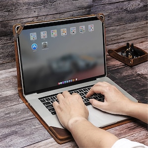 May include: A brown leather laptop sleeve with a flap closure. The sleeve is open and a laptop is inside. The laptop screen is on and shows a blurred image of a car. The laptop keyboard is visible and a person's hands are typing on the keyboard.