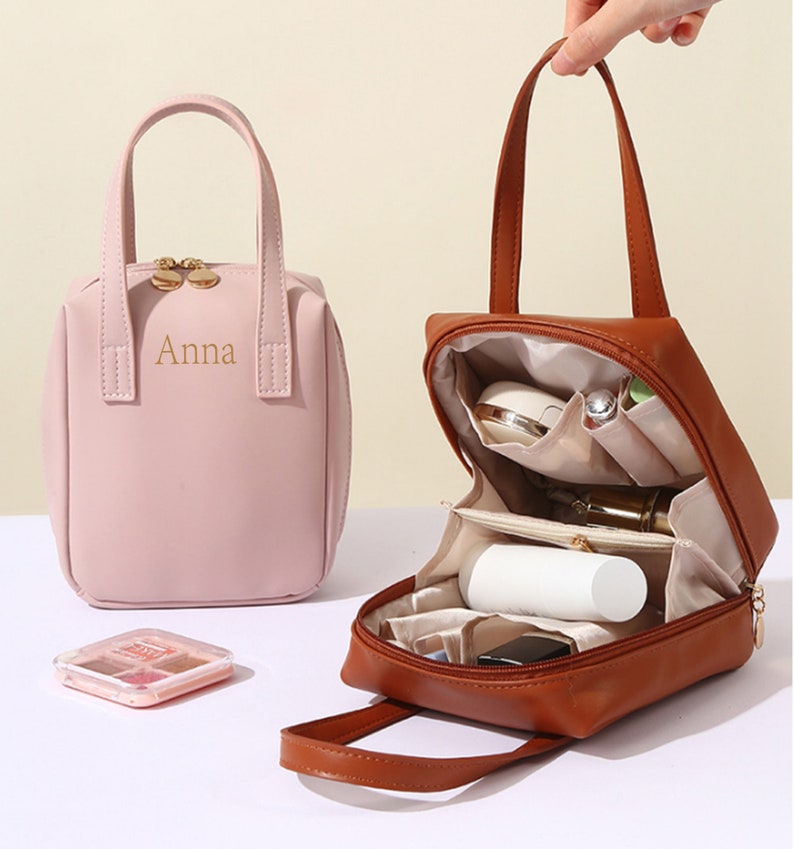 May include: Two small, square-shaped, zippered bags with handles. The bag on the left is pink and has the name "Anna" embroidered on the front. The bag on the right is brown and is open to show the interior compartments and pockets.