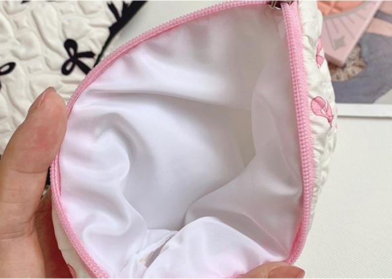 Op de afbeelding: Een witte cosmetische tas met een roze rits en interieur. De buitenkant van de tas heeft een gewatteerd patroon met roze bloemmotieven. De binnenkant is wit en lijkt gemaakt van een glad, zijdeachtig materiaal. De tas is open en onthult de binnenkant.