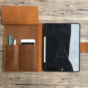 May include: A brown leather folio case with two slots for pens and a pocket for a phone. The case is open and a black tablet with a white stylus is inside.