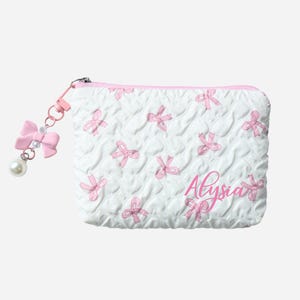 Op de afbeelding: Witte gewatteerde cosmetische tas met roze strikpatroon en de naam "Alysia" in roze script. Een roze strikbedel met een parel hangt aan de rits.