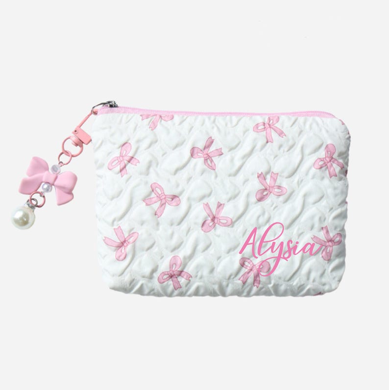 Op de afbeelding: Witte gewatteerde cosmetische tas met roze strikpatroon en de naam "Alysia" in roze script. Een roze strikbedel met een parel hangt aan de rits.