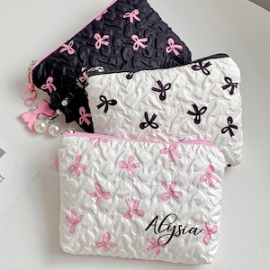 Op de afbeelding: Drie kleine, gewatteerde cosmetische tasjes. E&eacute;n is zwart met roze strikken, een andere is wit met zwarte strikken, en de derde is wit met roze strikken en de naam "Alysia". Elk tasje heeft een rits en een roze accent.