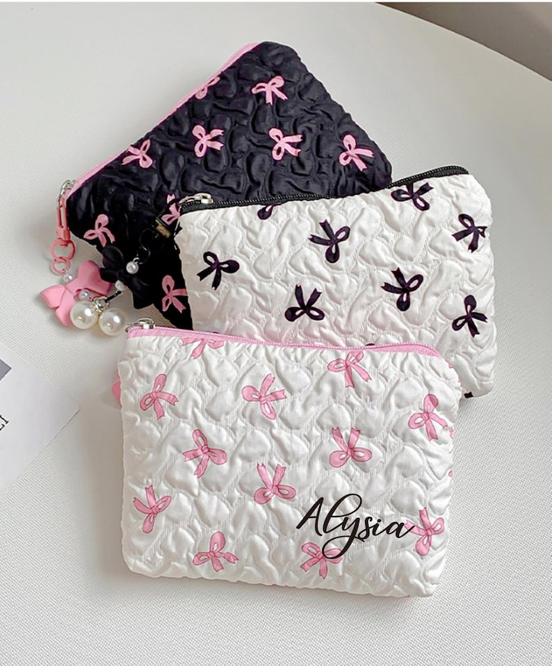 Op de afbeelding: Drie kleine, gewatteerde cosmetische tasjes. E&eacute;n is zwart met roze strikken, een andere is wit met zwarte strikken, en de derde is wit met roze strikken en de naam "Alysia". Elk tasje heeft een rits en een roze accent.
