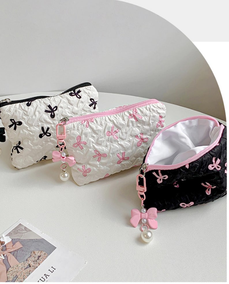 Op de afbeelding: Drie cosmetische tassen met strikpatronen. E&eacute;n is zwart met witte strikken, een andere is wit met roze strikken en de derde is zwart met roze strikken. Elke tas heeft een rits en een roze strikbedel met een parel.