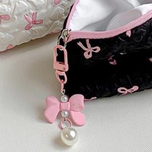Op de afbeelding: Zwarte make-uptas met roze strikaccenten en een bijpassende roze rits. Een roze strikbedel met parelaccenten hangt aan de rits. Een andere witte tas met roze strikaccenten staat op de achtergrond.