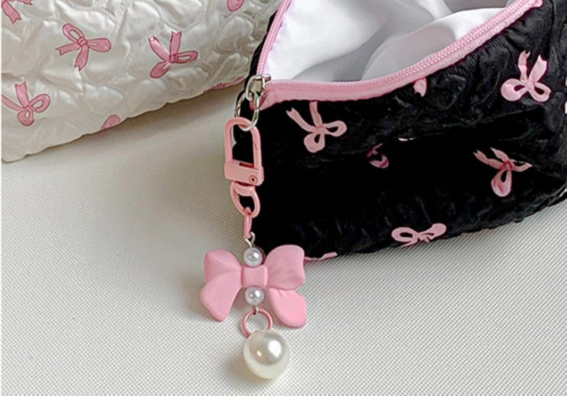 Op de afbeelding: Zwarte make-uptas met roze strikaccenten en een bijpassende roze rits. Een roze strikbedel met parelaccenten hangt aan de rits. Een andere witte tas met roze strikaccenten staat op de achtergrond.