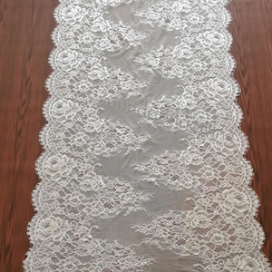 Op de afbeelding: Een witte kanten tafelloper met een bloemenpatroon. De loper is rechthoekig met geschulpte randen. Het kantontwerp heeft gedetailleerde bloemmotieven en een delicate, open weefsel, perfect om een vleugje elegantie aan elke tafelsetting toe te voegen.