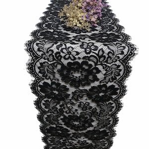 Op de afbeelding: Zwarte kanten tafelloper met bloemen- en bladerpatronen. De loper is bovenaan versierd met gedroogde bloemen. Het ingewikkelde ontwerp en de delicate textuur maken het geschikt voor diverse evenementen en woondecoratie.