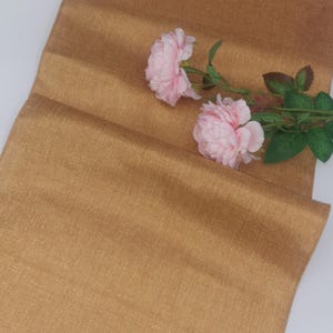 Può includere: Un runner da tavolo dorato con un leggero luccichio, che presenta una texture delicata. Due fiori rosa sono posizionati sul runner.