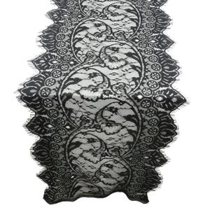 May include: Black lace table runner with a detailed floral and scroll design. The runner has a scalloped edge with a fringe. The intricate pattern is visible against a white background, highlighting the delicate lace.