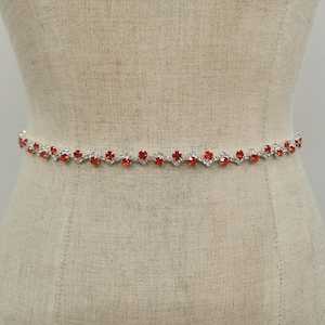 Könnte beinhalten: Ein silberfarbener Gürtel mit einem sich wiederholenden Muster aus roten herzförmigen Edelsteinen und klaren Kristallen. Der Gürtel wird auf einer Schaufensterpuppe präsentiert, die sein zartes Design zeigt.