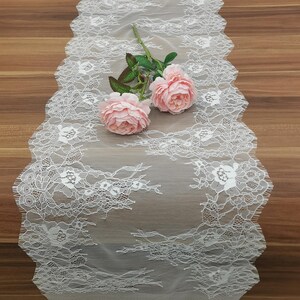 May include: White lace table runner with a scalloped edge, adorned with floral patterns. Two pink roses with green leaves are placed on the runner, creating a delicate and romantic aesthetic. The runner is on a wooden surface.