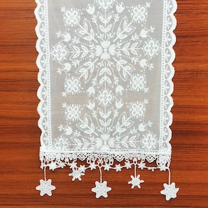 May include: White lace table runner with a floral and leaf pattern. The runner has a scalloped edge and decorative star-shaped accents hanging from the bottom. The runner is displayed on a wooden surface.