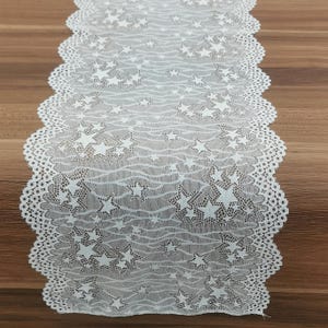 May include: White lace table runner with a star pattern. The runner has a scalloped edge and is placed on a wooden surface. The star design is evenly distributed across the fabric, creating a delicate and elegant look.