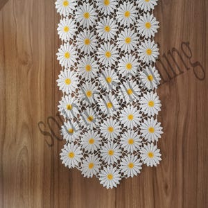 May include: White lace table runner featuring a daisy pattern with yellow centers. The runner is placed on a wooden surface. The daisy design is repeated throughout the length of the runner, creating a delicate and floral aesthetic.