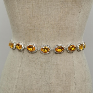 May include: A silver belt with oval-shaped amber-colored gemstones, each surrounded by smaller clear crystals. The belt is displayed on a mannequin, showcasing its design and potential fit.