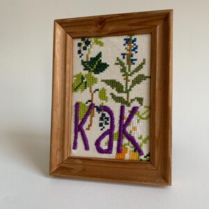 Puede incluir: Un bordado de punto de cruz enmarcado con un diseño floral y la palabra "kak" en letras moradas.