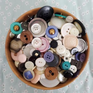 May include: A round wooden bowl filled with a large collection of assorted sewing buttons in various sizes, shapes, and colors, including white, black, blue, pink, brown, and teal, featuring both two-hole and four-hole designs.