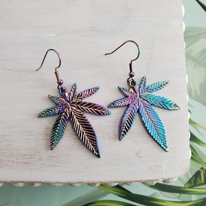 May include: A pair of iridescent dangle earrings featuring a marijuana leaf design. The earrings are made of metal and have a silver hook.
