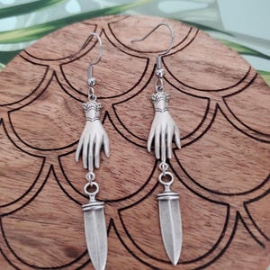 May include: A pair of silver earrings featuring a hand holding a dagger charm. The earrings are on a wooden surface with a decorative pattern.