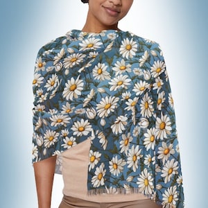 Puede incluir: Una mujer lleva una bufanda floral azul y blanca con estampado de margaritas. La bufanda está drapeada alrededor de su cuello y hombros.