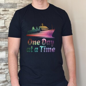 Puede incluir: Camiseta negra con un diseño colorido de crucero. El barco está representado en tonos rosas, morados y verdes. Debajo, el texto "One Day at a Time" está escrito en una fuente de gradiente arcoíris. Es una prenda de vestir informal.