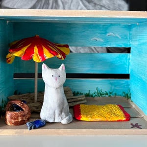 May include: A wooden box diorama featuring a beach scene. A white cat figurine sits near a yellow and red beach umbrella. A yellow and red beach towel and a brown basket are also present. The background is painted blue with seagull details.