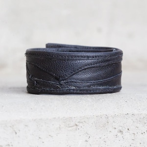 Puede incluir: Una pulsera de cuero negro con una superficie texturizada y detalles de costura visibles a lo largo de los bordes superior e inferior. La banda tiene un diseño curvo y superpuesto con un patrón de costura en forma de V en el centro exterior.