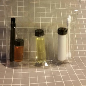 May include: A clear plastic bag with three small bottles and two cotton swabs. The bottles are filled with clear liquids, one is amber colored, one is yellow, and one is white. The bag is on a gray grid background.