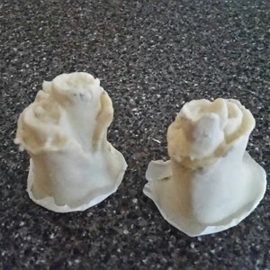 May include: Two white ceramic cake toppers shaped like roses. The toppers are in the shape of a single rose with a stem and leaves.