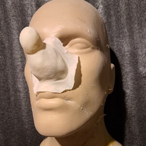 May include: A close-up of a prosthetic nose made of silicone. The nose is attached to a mannequin head with a piece of white paper.