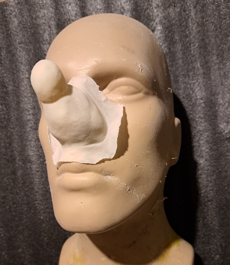 May include: A close-up of a prosthetic nose made of silicone. The nose is attached to a mannequin head with a piece of white paper.