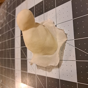 May include: A white plaster mold of a bird with a round head and a slightly curved body. The mold is sitting on a black and white grid surface.