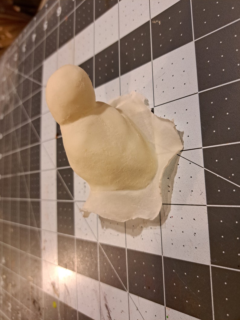 May include: A white plaster mold of a bird with a round head and a slightly curved body. The mold is sitting on a black and white grid surface.