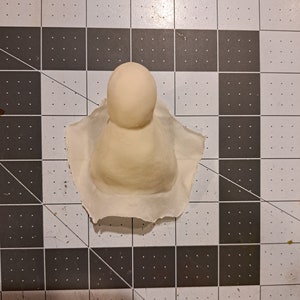 May include: A white clay sculpture of a person with a round head and a cone-shaped body.
