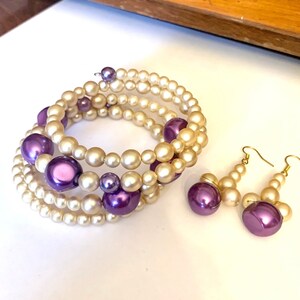 May include: A set of pearl and purple bead jewelry featuring a multi-strand bracelet and a pair of dangle earrings.