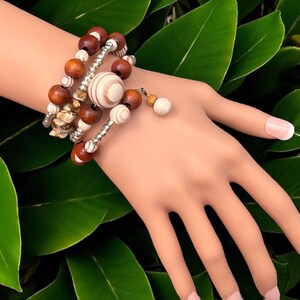 May include: A multi-strand bracelet featuring a mix of wooden beads in shades of brown and cream, interspersed with silver-toned metal accents. The bracelet includes a large, striped wooden bead and a small dangling charm. The jewelry is displayed against a backdrop of green leaves.