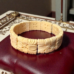 Könnte beinhalten: Elfenbeinfarbenes Armband mit geschnitzten Blumen- und geometrischen Mustern. Das Armband besteht aus rechteckigen Segmenten, mit Blumenmustern und kreuzförmigen Designs. Es liegt auf einer weinroten Oberfläche.