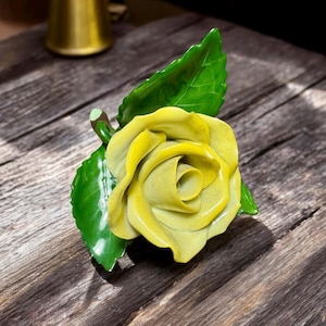 Puede incluir: Una rosa de cerámica amarilla con hojas verdes. La rosa es una representación realista de una flor.