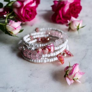 May include: A white beaded bracelet with pink and white beads and pink shell accents. The bracelet is layered and has a delicate, feminine look.