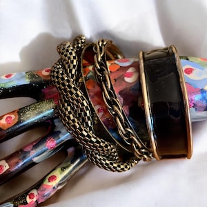 Pode incluir: Um conjunto de quatro pulseiras douradas, incluindo uma pulseira de corrente, uma pulseira tecida e duas pulseiras largas. Uma pulseira é preta.